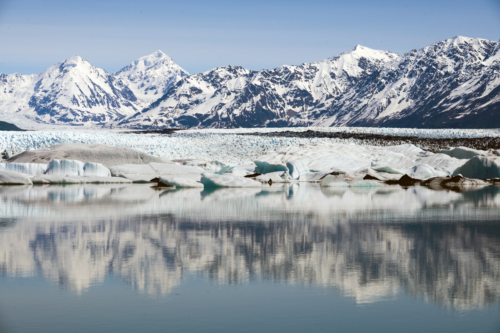 Snow-covered peaks and a glacier reflected in still Alaskan water.