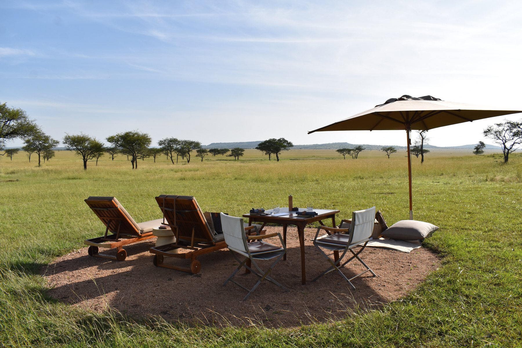 Safari loungers and a dining table set under an umbrella on the open savannah.